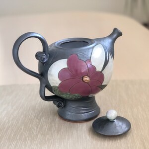 May include: A black ceramic teapot with a floral design. The teapot has a lid and a handle. The floral design features a large pink flower with a white center and green leaves.