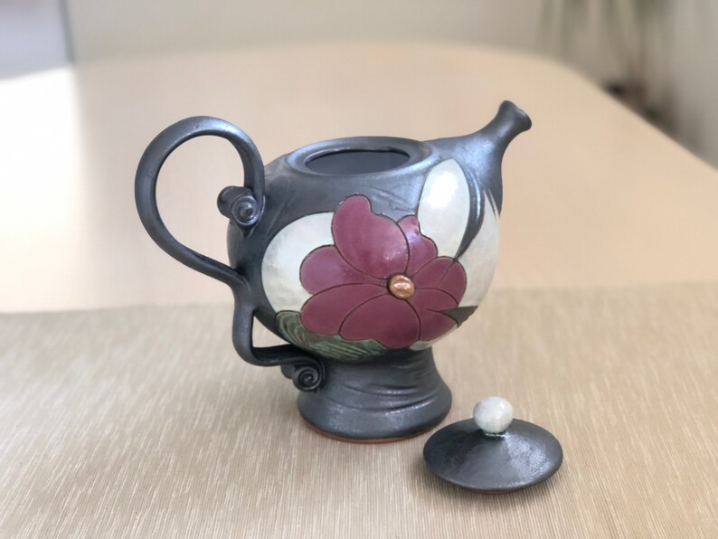 May include: A black ceramic teapot with a floral design. The teapot has a lid and a handle. The floral design features a large pink flower with a white center and green leaves.