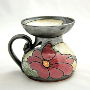 Op de afbeelding: Een keramische mok met een grijze glazuur en een bloemenontwerp. De mok heeft een handvat en een brede, ondiepe bovenkant. De bloem is roze met een gele kern en groene bladeren.