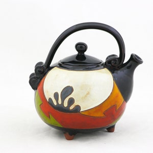 Könnte beinhalten: Eine schwarze Teekanne aus Keramik mit einem geometrischen Design in Weiß, Orange, Rot und Grün. Die Teekanne hat einen schwarzen Henkel und Ausguss und steht auf drei kleinen Füßen.