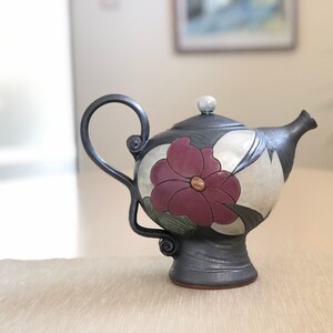May include: A black ceramic teapot with a floral design. The teapot has a single flower with a pink center and green leaves. The teapot has a curved spout and a handle.