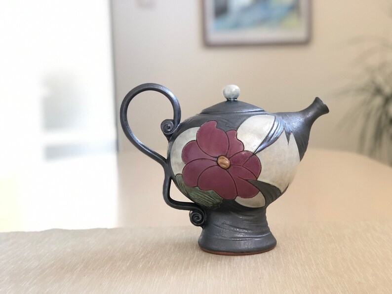 May include: A black ceramic teapot with a floral design. The teapot has a single flower with a pink center and green leaves. The teapot has a curved spout and a handle.