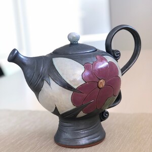 May include: A black ceramic teapot with a floral design. The teapot has a purple flower with a yellow center and black leaves. The teapot has a round body and a spout on the side. The teapot has a handle on the top and a base with a round foot.