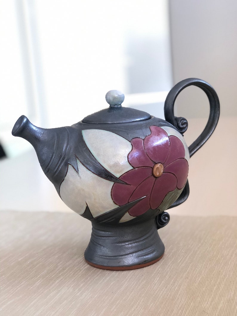May include: A black ceramic teapot with a floral design. The teapot has a purple flower with a yellow center and black leaves. The teapot has a round body and a spout on the side. The teapot has a handle on the top and a base with a round foot.