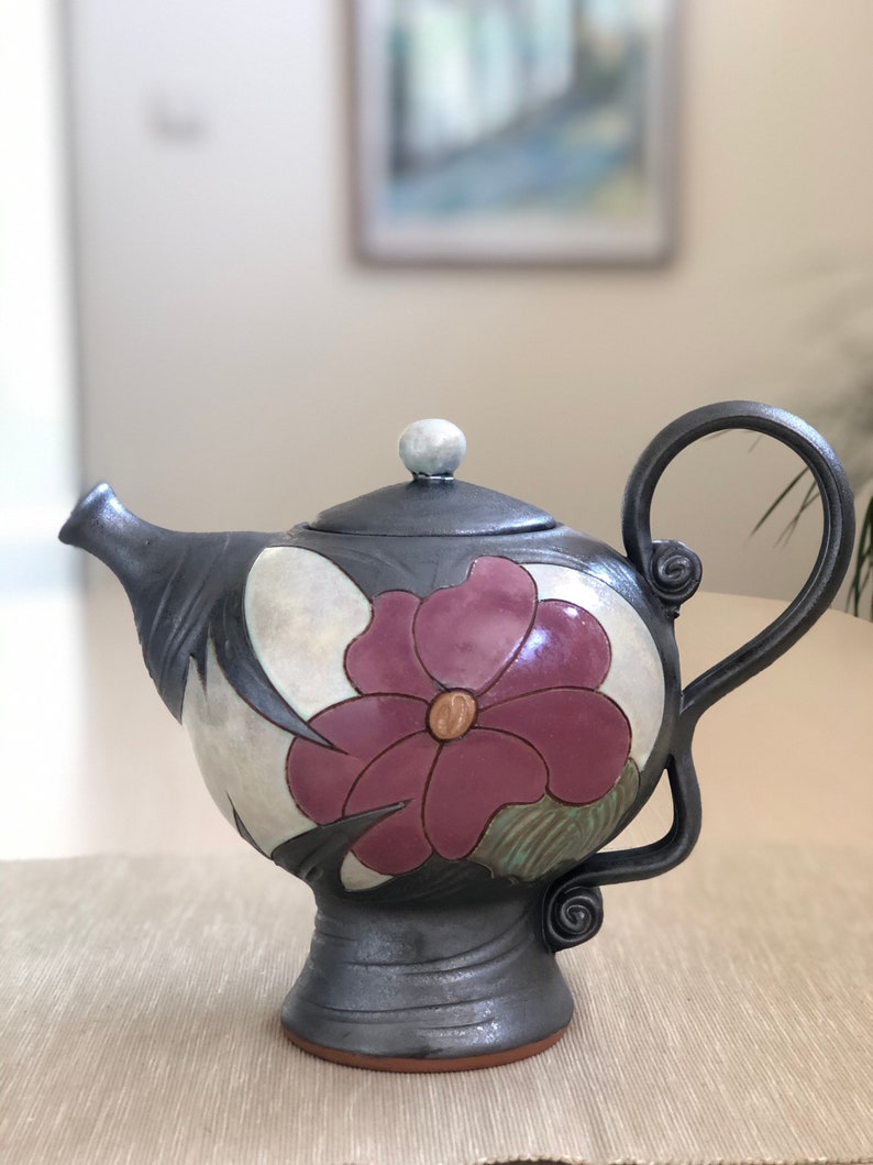 May include: A black ceramic teapot with a floral design. The teapot has a round body with a single handle and a lid with a knob. The floral design features a large pink flower with a yellow center and green leaves.
