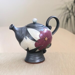 May include: A black ceramic teapot with a floral design. The teapot has a white background with a large purple flower and black leaves. The teapot has a round body and a small spout.