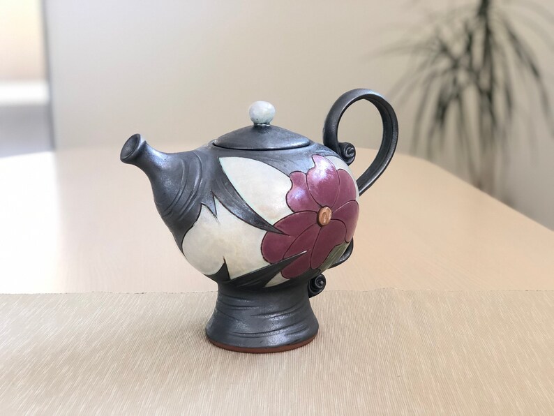 May include: A black ceramic teapot with a floral design. The teapot has a white background with a large purple flower and black leaves. The teapot has a round body and a small spout.