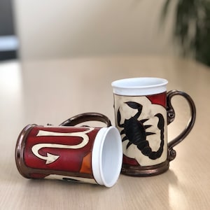 Può includere: Due tazze in ceramica con segni zodiacali. Una tazza presenta uno sfondo rosso e arancione con un simbolo del Sagittario dorato. L'altra tazza ha uno sfondo bianco con uno scorpione nero, il segno zodiacale dello Scorpione.
