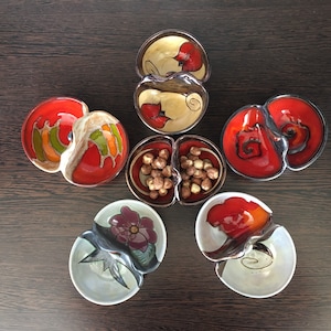 May include: Five ceramic serving dishes with two compartments each. The dishes are decorated with red, orange, yellow, green, brown, and black paint. The dishes are arranged in a star shape on a dark brown wooden surface.