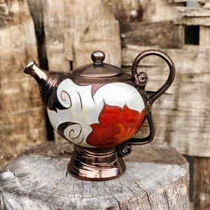 Peut inclure: Une théière en céramique peinte à la main avec un glaçage cuivré. La théière a un fond blanc avec un motif floral rouge et orange. La théière a un corps rond et un bec courbé.