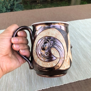 May include: A brown ceramic mug with a white interior and a decorative design of swirling lines and shapes in brown and cream.