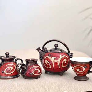Può includere: Un set da tè in ceramica rosso e marrone con una teiera, un creamer, una zuccheriera e una tazza da tè. Il set presenta un design a spirale bianco.