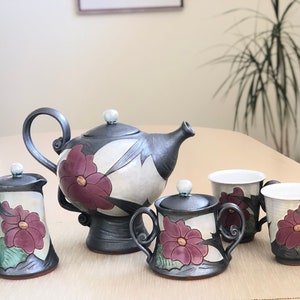May include: A ceramic tea set with a teapot, creamer, sugar bowl, and two cups. The set is decorated with a floral design in shades of white, black, and purple.
