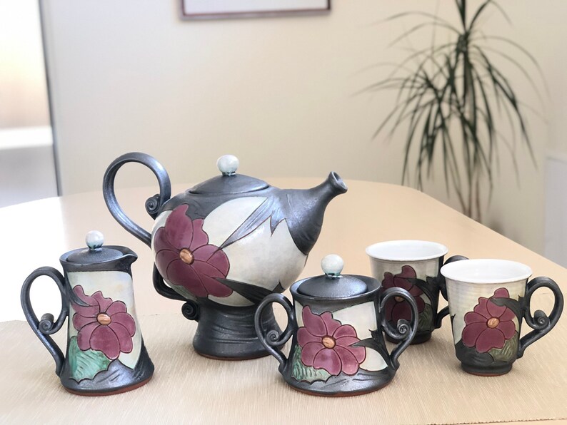 May include: A ceramic tea set with a teapot, creamer, sugar bowl, and two cups. The set is decorated with a floral design in shades of white, black, and purple.