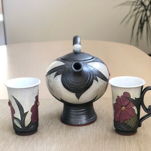 May include: A ceramic teapot and two mugs with floral designs. The teapot is black and white with a floral pattern. The mugs are white with a floral pattern in shades of purple and green. The teapot has a spout and a handle. The mugs have handles.