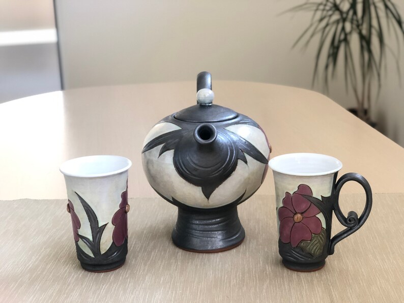 May include: A ceramic teapot and two mugs with floral designs. The teapot is black and white with a floral pattern. The mugs are white with a floral pattern in shades of purple and green. The teapot has a spout and a handle. The mugs have handles.
