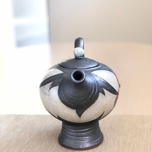 May include: A black and white ceramic teapot with a floral design. The teapot has a round body and a short spout. The lid has a small knob on top.