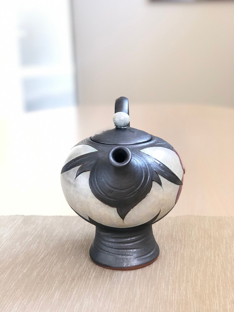 May include: A black and white ceramic teapot with a floral design. The teapot has a round body and a short spout. The lid has a small knob on top.