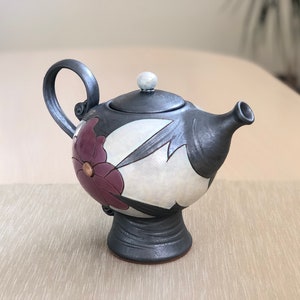 May include: A black and white ceramic teapot with a floral design. The teapot has a round body with a spout and a handle. The teapot is sitting on a table.