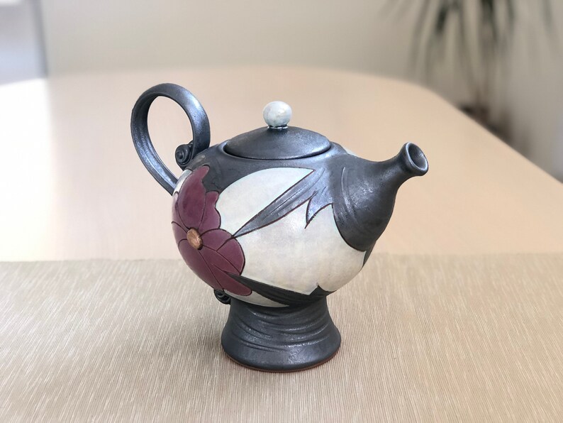 May include: A black and white ceramic teapot with a floral design. The teapot has a round body with a spout and a handle. The teapot is sitting on a table.