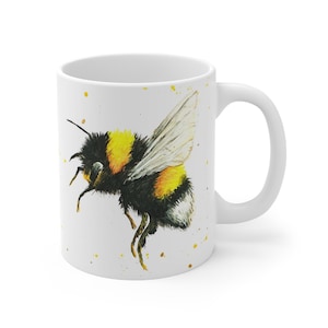 May include: White ceramic coffee mug featuring a watercolor illustration of a bumblebee in flight. The bee has black and yellow stripes and detailed wings. Yellow paint splatters accent the mug's surface, creating a whimsical design.