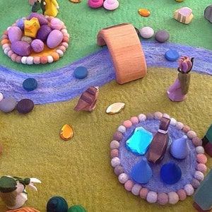 May include: A felt play mat with a blue river, a wooden bridge, and various colorful felt figures and objects. The figures include a bird, a duck, a flower, and a person. The objects include a small wooden house, a small wooden tree, and a small wooden flower.