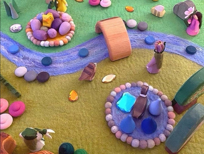 May include: A felt play mat with a blue river, a wooden bridge, and various colorful felt figures and objects. The figures include a bird, a duck, a flower, and a person. The objects include a small wooden house, a small wooden tree, and a small wooden flower.