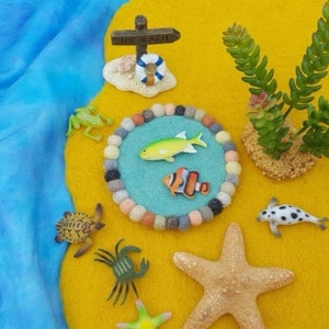 May include: A felt play mat with a blue circle in the center. The circle has a yellow fish, an orange and white fish, and a variety of colorful felt balls around the edge. There are also a green frog, a turtle, a crab, a starfish, and a black and white fish on the mat. The mat is on a yellow background with a sign that says "The Beach".