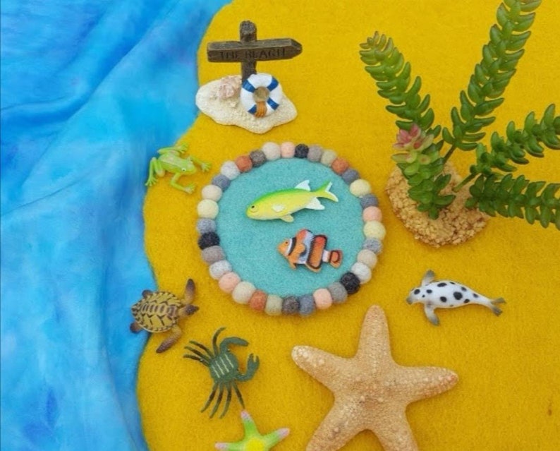May include: A felt play mat with a blue circle in the center. The circle has a yellow fish, an orange and white fish, and a variety of colorful felt balls around the edge. There are also a green frog, a turtle, a crab, a starfish, and a black and white fish on the mat. The mat is on a yellow background with a sign that says "The Beach".