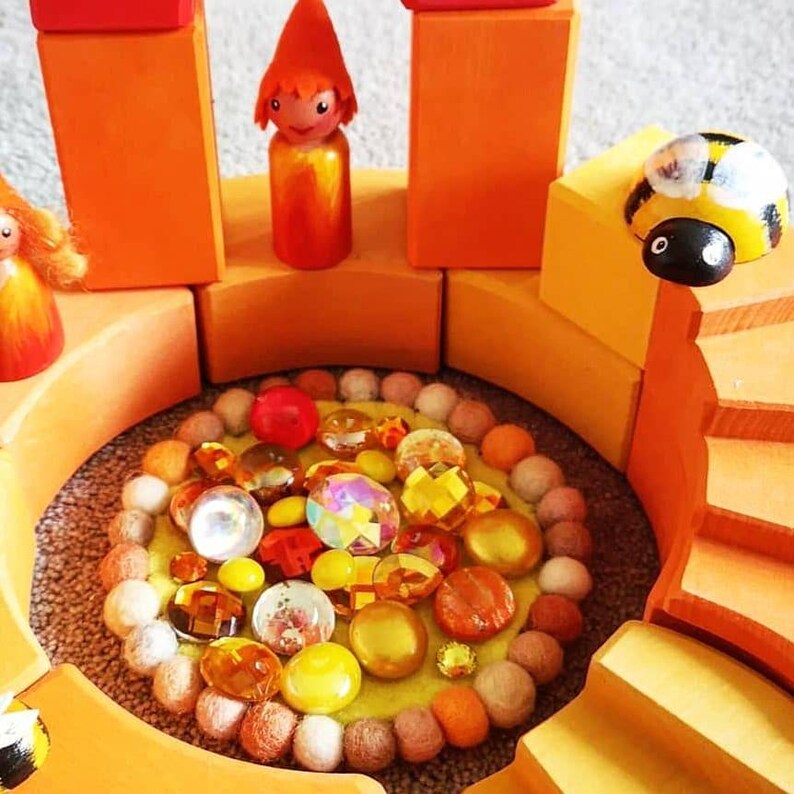 May include: A wooden toy castle with orange and red walls, a small yellow and orange gnome figure, and a bee figure. The castle has a circular opening with a yellow felt base and a variety of colorful gems and beads.