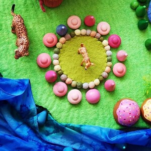 May include: A green felt play mat with a circle of pink felt balls and a deer figurine in the center. There are also a leopard figurine, a blue fabric, and a few trees on the mat.