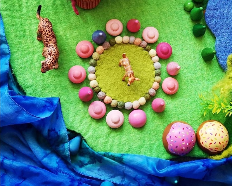 May include: A green felt play mat with a circle of pink felt balls and a deer figurine in the center. There are also a leopard figurine, a blue fabric, and a few trees on the mat.