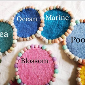 May include: Six colorful felt coasters with pom-pom trim. The coasters are labeled "Sea", "Ocean", "Marine", "Pool", "Blossom", and an unlabeled yellow coaster.