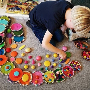 May include: A colorful felt circle garland with a variety of wooden toys, including small balls, cones, and other shapes. The garland is arranged in a spiral pattern on a light gray carpet.