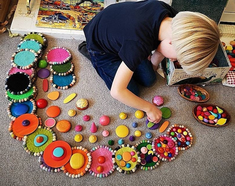 May include: A colorful felt circle garland with a variety of wooden toys, including small balls, cones, and other shapes. The garland is arranged in a spiral pattern on a light gray carpet.