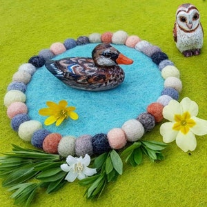 May include: A blue felt circle with a brown and white duck figurine in the center. The circle is surrounded by a ring of colorful felt balls. There are two yellow flowers and green leaves in the foreground.