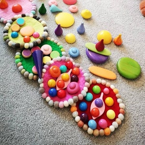 May include: A set of colorful felt circles with various wooden shapes and beads arranged in a circular pattern. The circles are decorated with different colors and patterns, including pink, green, yellow, and red. The wooden shapes include small circles, squares, and triangles.