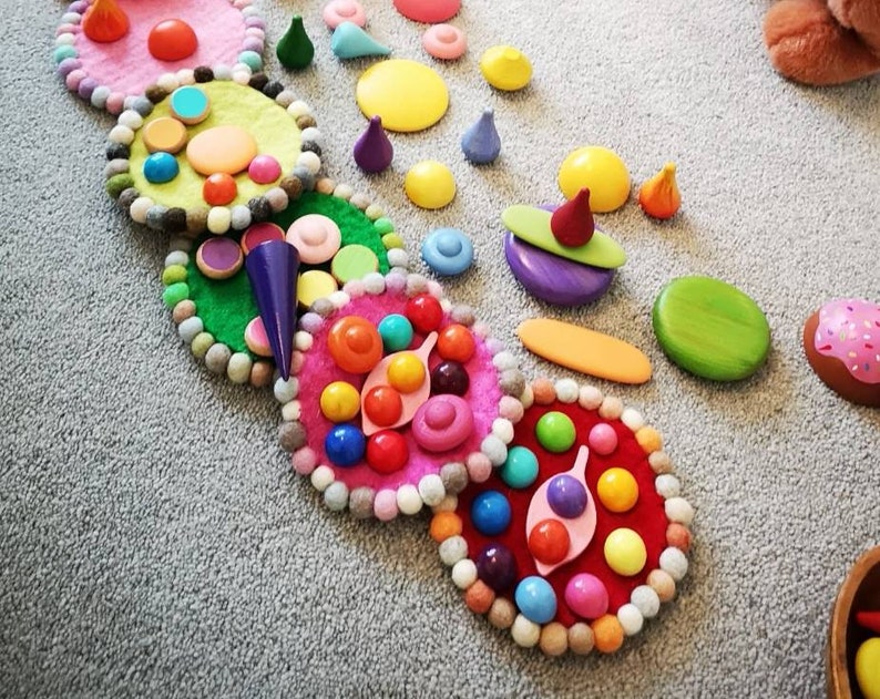 May include: A set of colorful felt circles with various wooden shapes and beads arranged in a circular pattern. The circles are decorated with different colors and patterns, including pink, green, yellow, and red. The wooden shapes include small circles, squares, and triangles.