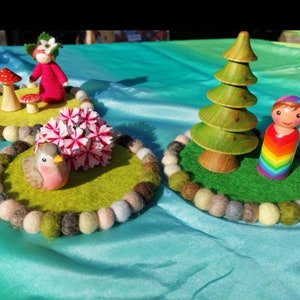 May include: Three felt play mats with a doll, a bird, and a tree. The mats are green and have a circle of colorful felt balls around the edge. The doll is wearing a rainbow dress and has brown hair. The bird is pink and white. The tree is green and brown.