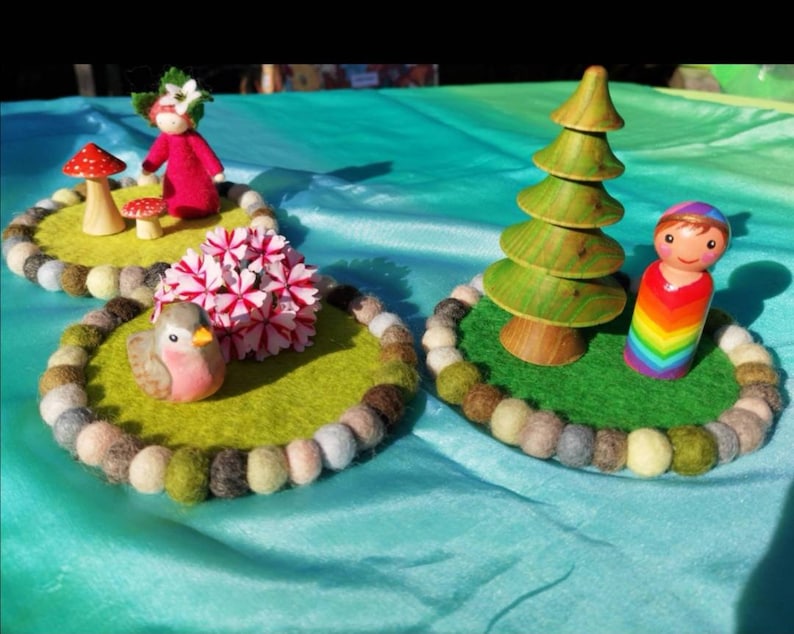 May include: Three felt play mats with a doll, a bird, and a tree. The mats are green and have a circle of colorful felt balls around the edge. The doll is wearing a rainbow dress and has brown hair. The bird is pink and white. The tree is green and brown.