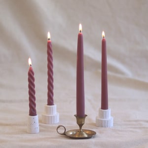 May include: Three pink candles on different candle holders. The candles are lit and the holders are white with a speckled finish. The center candle is on a gold metal candlestick.