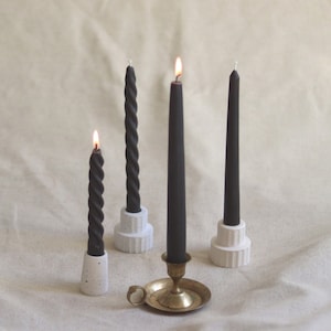 May include: An assortment of black taper candles, some lit, in various holders. Two twisted candles sit in white concrete holders, while a straight candle rests in a brass candlestick. The candles are on a neutral-toned surface.