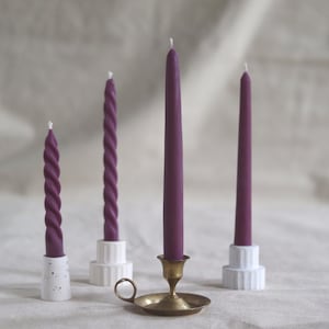 May include: Three purple candles on different holders. The candle on the left is a twisted candle on a white holder with black speckles. The middle candle is a twisted candle on a white holder with a ridged design. The candle on the right is a straight candle on a white holder with a ridged design. The candle in the middle is on a gold candlestick.