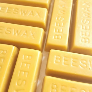 May include: A close-up shot of several rectangular beeswax bars. The bars are a warm yellow hue and have the word "BEESWAX" embossed on them. They are arranged in a grid, indicating a large quantity of the product.