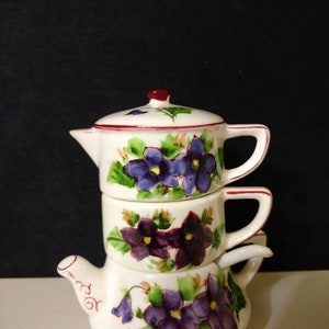 May include: A set of three white ceramic teapots with purple and green floral designs. The teapots are stacked on top of each other, with the smallest teapot on top.