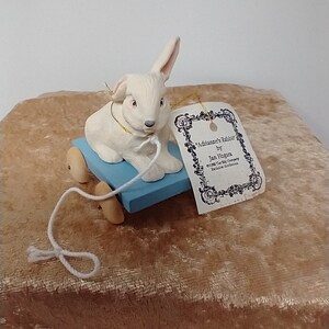 May include: White porcelain rabbit figurine on a blue wooden wagon with wheels. The rabbit is pulling a white string attached to the wagon. The figurine has a tag that reads "Adrienne's Rabbit by Jan  Hagara Exclusive Edition Made in Germany".