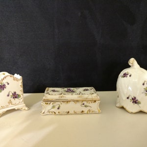 May include: A set of three vintage porcelain trinket boxes with a floral design. The boxes are decorated with purple flowers and gold accents. The boxes are in the shape of a square, a rectangle, and a bell.
