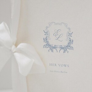 May include: A wedding vow booklet with a blue floral design and the text "HER VOWS". The booklet is tied with a white satin ribbon. The design includes the initials "E L" inside a decorative frame.