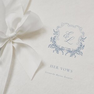May include: A white wedding vow book with a light blue floral design and the text "HER VOWS". A white ribbon is tied in a bow on the left side of the book. The book is made of textured paper.
