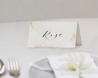 Könnte beinhalten: Weiße Tischkarte mit goldenen Akzenten und dem Namen "Rose" in schwarzer Kalligraphie.